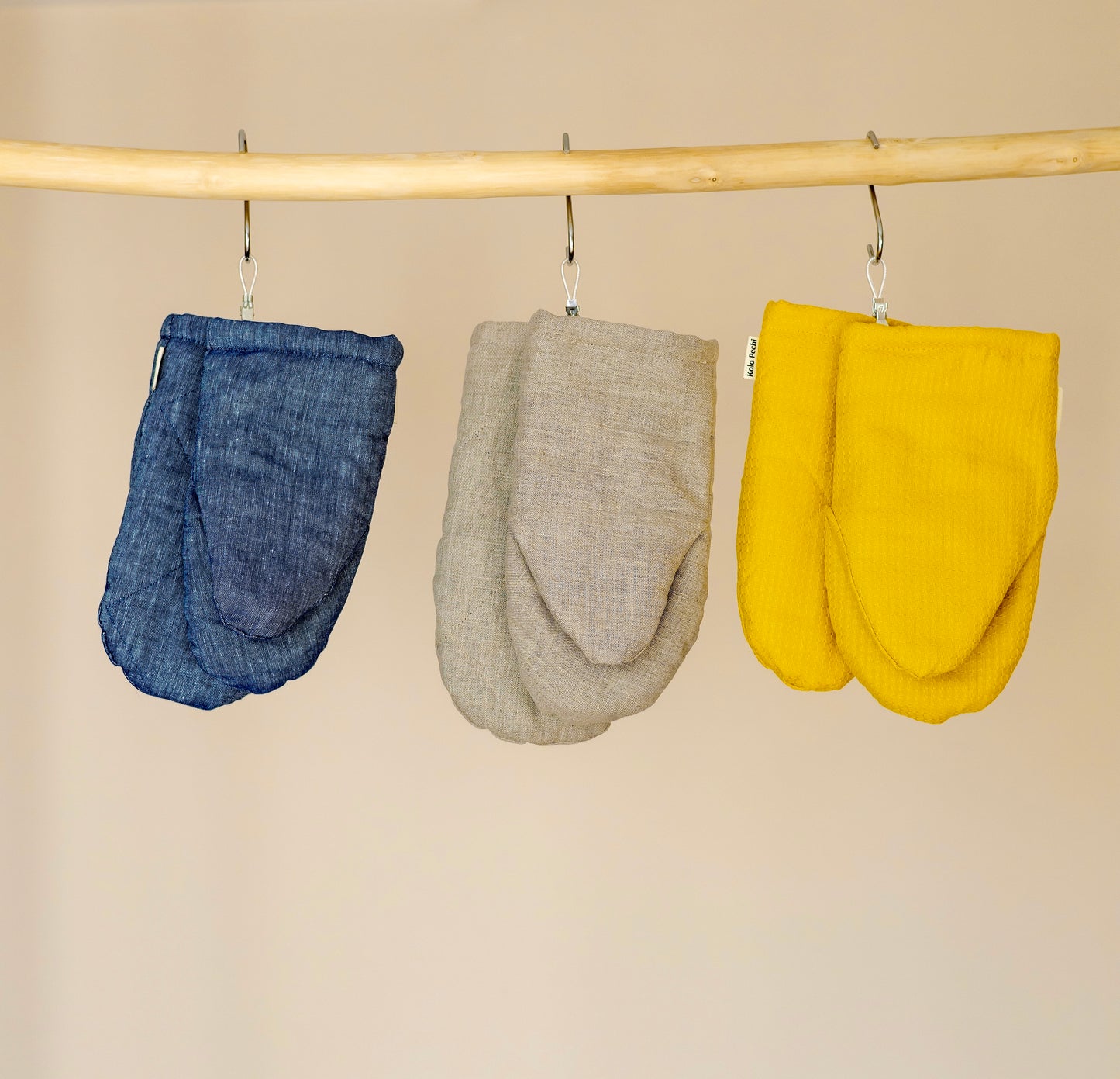Three linen and hemp oven mitts by Kolo Pechi in blue, beige, and yellow hanging on a rod against a beige background.