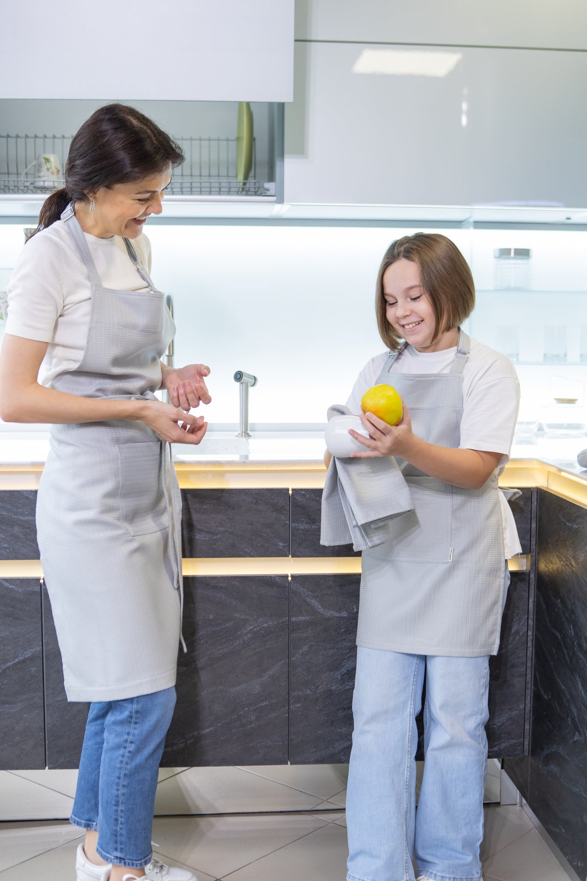 A woman and a young girl wearing matching light gray Kolo Pechi aprons stand in a modern kitchen. The girl holds a yellow lemon wrapped in a gray towel.
