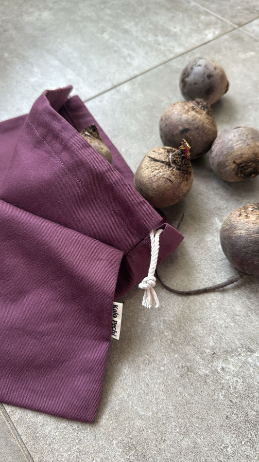 A purple reusable Kolo Pechi fabric bag from The Borscht Collection with a white drawstring closure, shown on a gray tiled floor next to several beets.