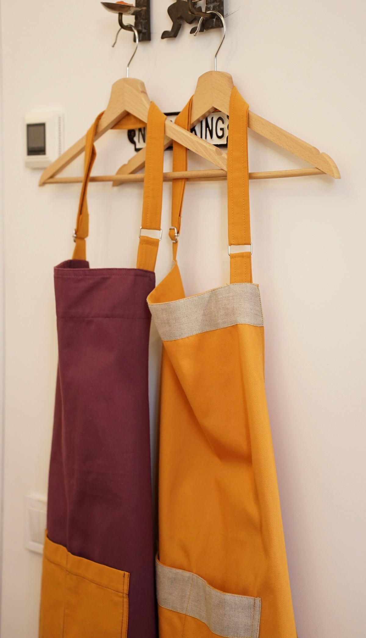 Two fabric Kolo Pechi Bib aprons in size Uni hanging on wooden hangers against a white wall. The apron on the left is solid deep purple, and the apron on the right is mustard yellow with a light gray band and pocket.