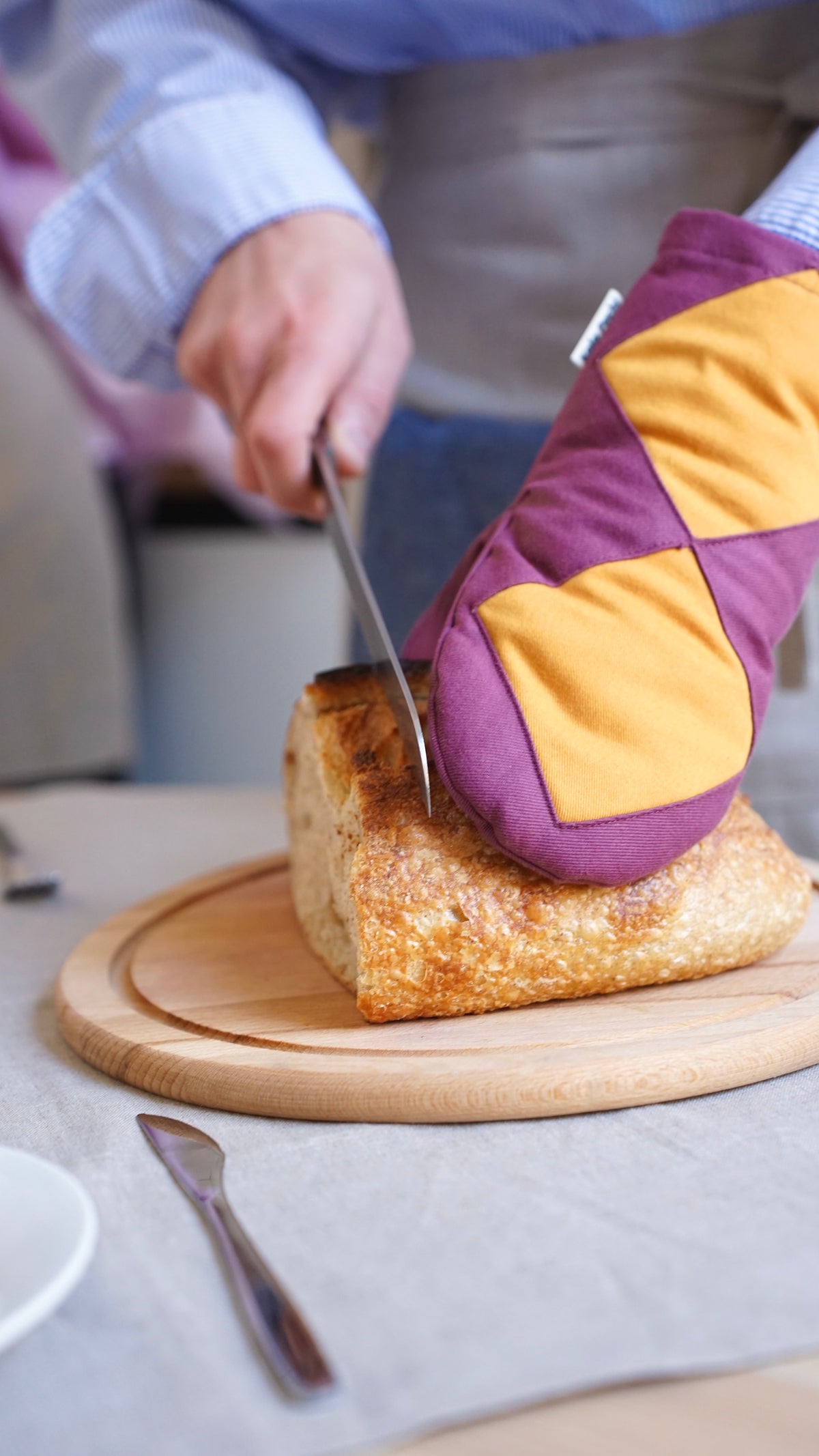 A close-up lifestyle shot of a person slicing a crusty loaf of bread on a wooden board, wearing a purple and mustard yellow Kolo Pechi oven mitt from the Borscht Collection for protection.
