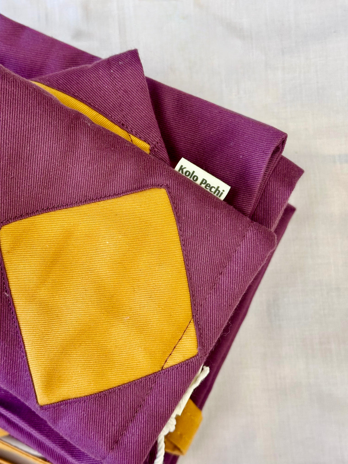 A folded purple fabric Oven Mitt From The Kolo Pechi Borscht Collection with a large mustard yellow patch and a visible tag reading 'kolo pechi', photographed on a light neutral fabric background.