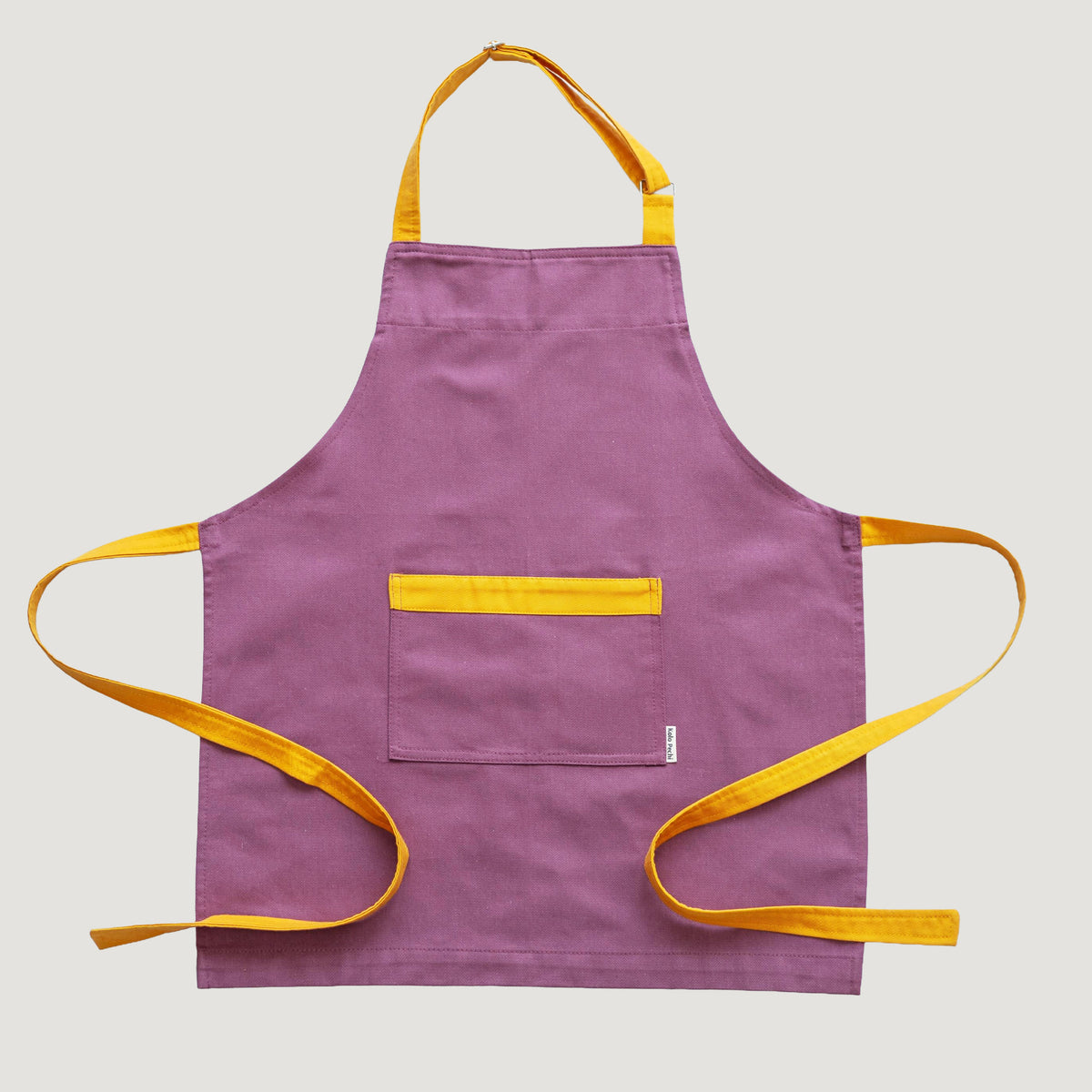 A purple Kolo Pechi Bib apron from The Borscht Collection with mustard yellow straps, neck loop, and pocket trim, laid flat against a light gray background.