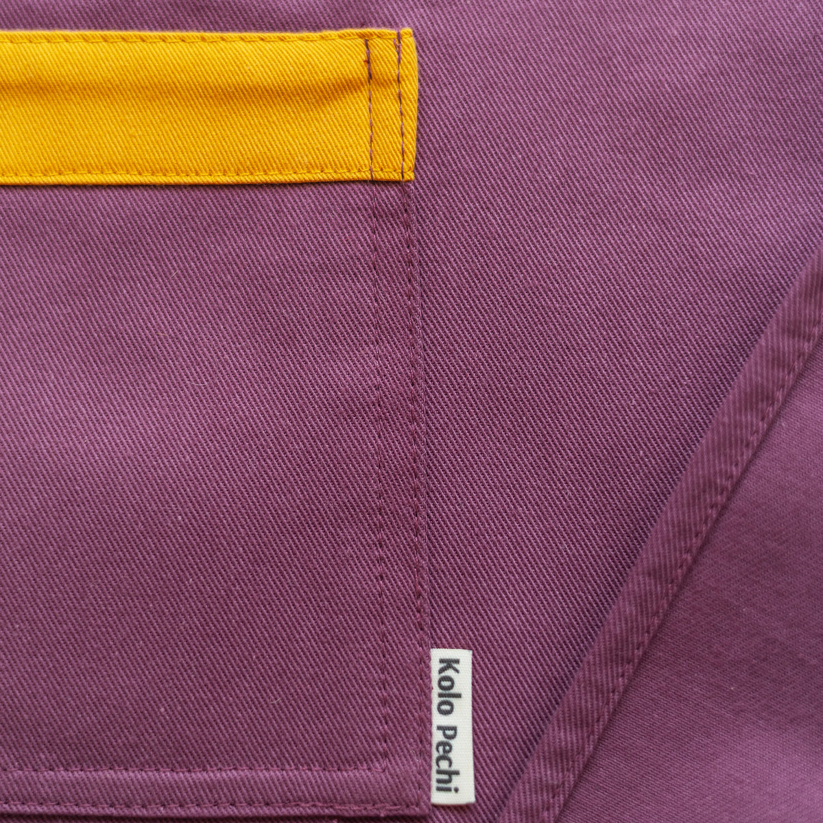 Close-up of a purple Kolo Pechi Bib Apron from the Borscht Collection  with a mustard yellow pocket trim and a small white brand tag reading Kolo Pechi.