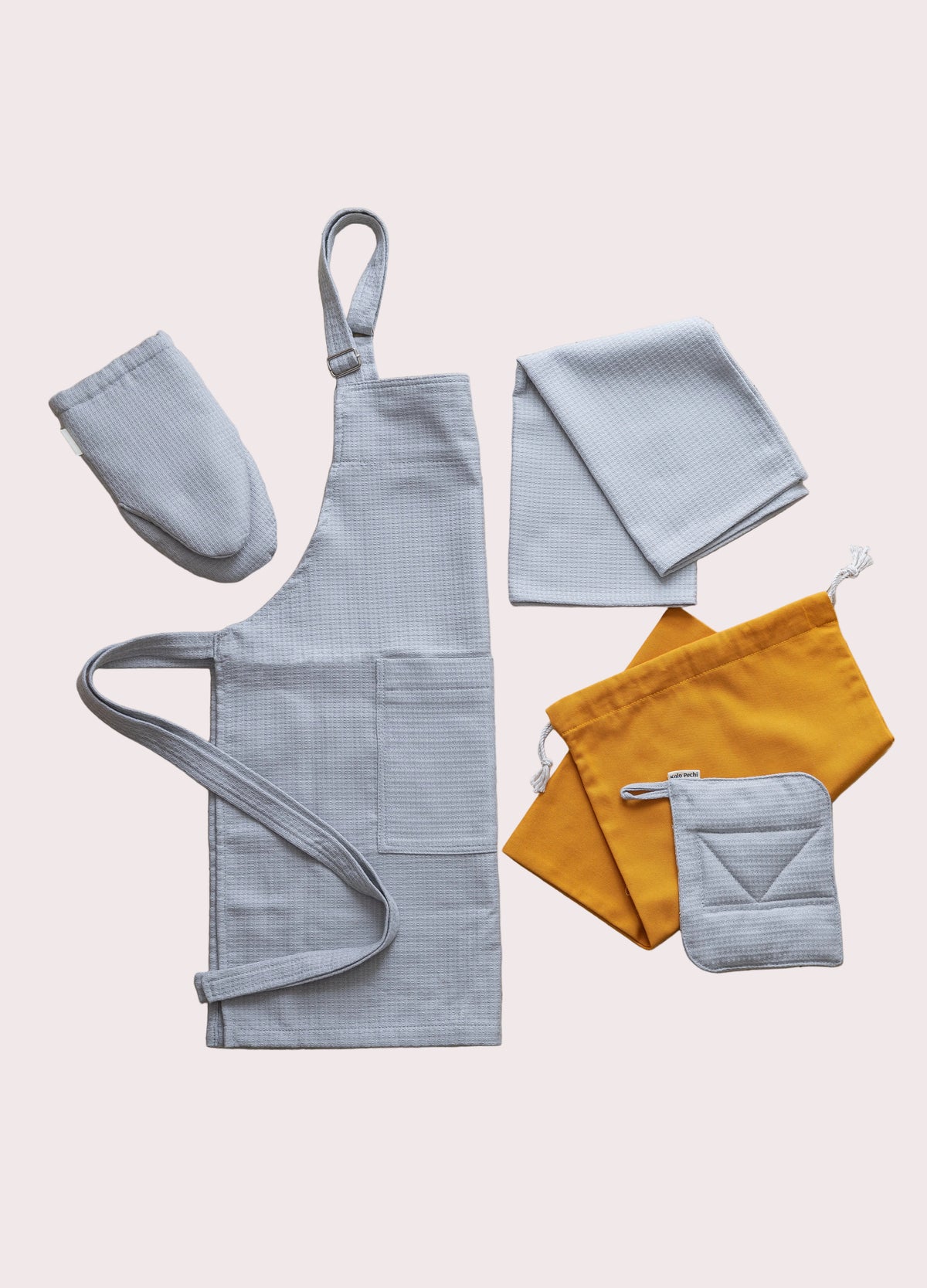 A flat-lay of The Kolo Pechi  kitchen textile set from the cotton Lviv Sky collection. The set includes a light gray waffle-weave apron, matching bib, oven mitt, pot holder, kitchen towel and drawstring grocery bag  in mustard yellow.