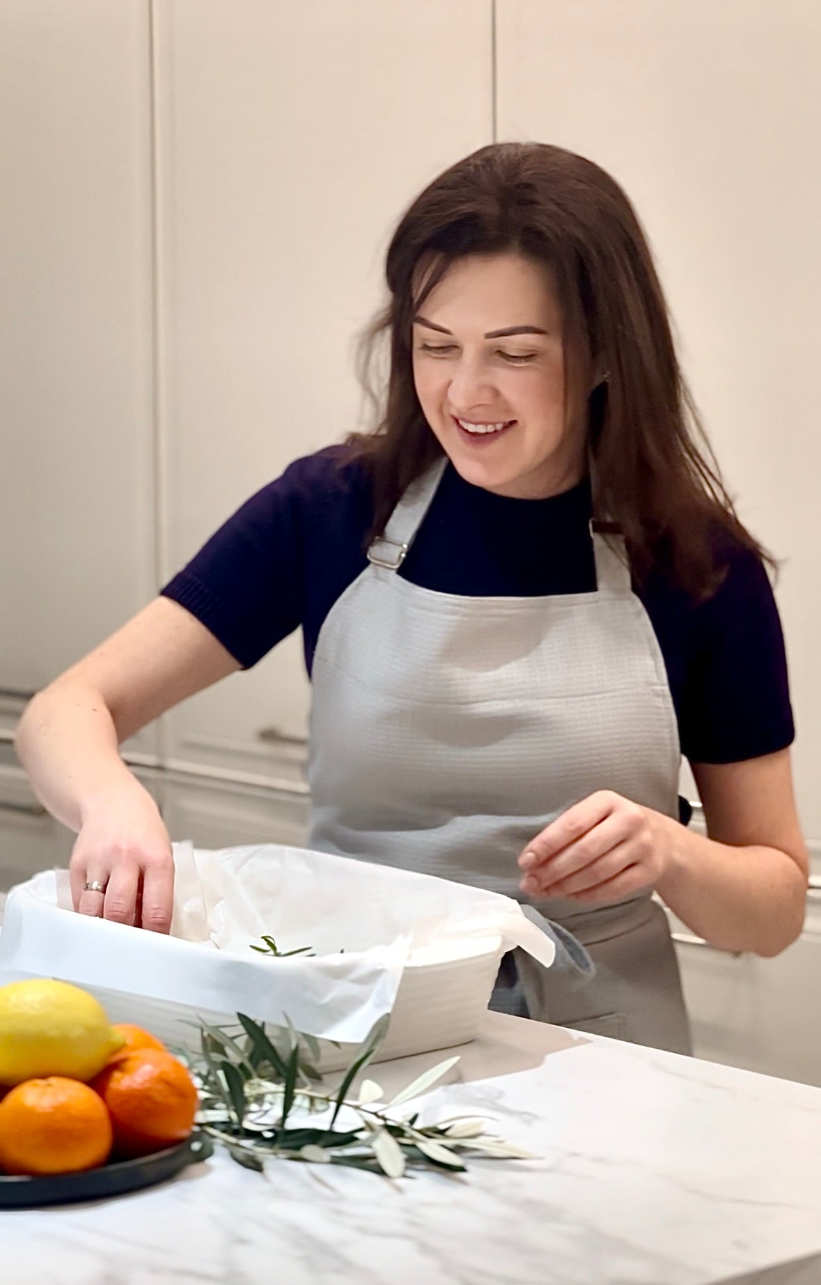 A woman wearing a grey cotton Kolo Pechi apron over a dark navy t-shirt is smiling while preparing food in a kitchen setting. In the foreground, a marble countertop holds fresh green herbs and a plate with lemons and oranges.