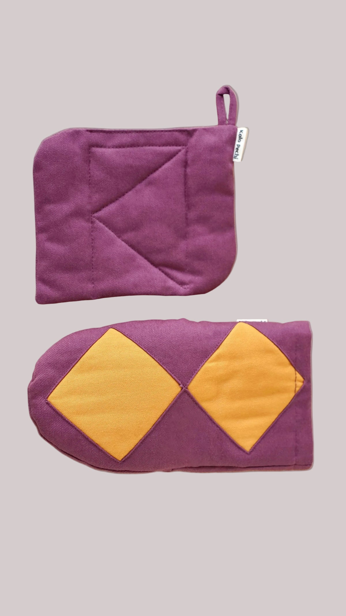 A pair of purple quilted Kolo Pechi oven mitts from The Borscht Collection on a light wood background. The top mitt is solid purple with a small white tag, and the bottom mitt is purple with two mustard yellow diamond-shaped patches.