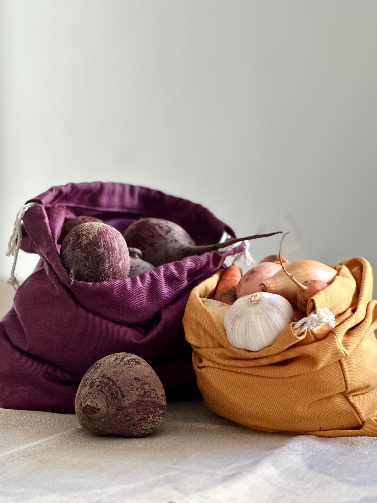 Two fabric produce bags on a light surface against a soft gray background. One bag is deep purple and contains beets, while the other is mustard yellow and contains onions, garlic, and root vegetables.