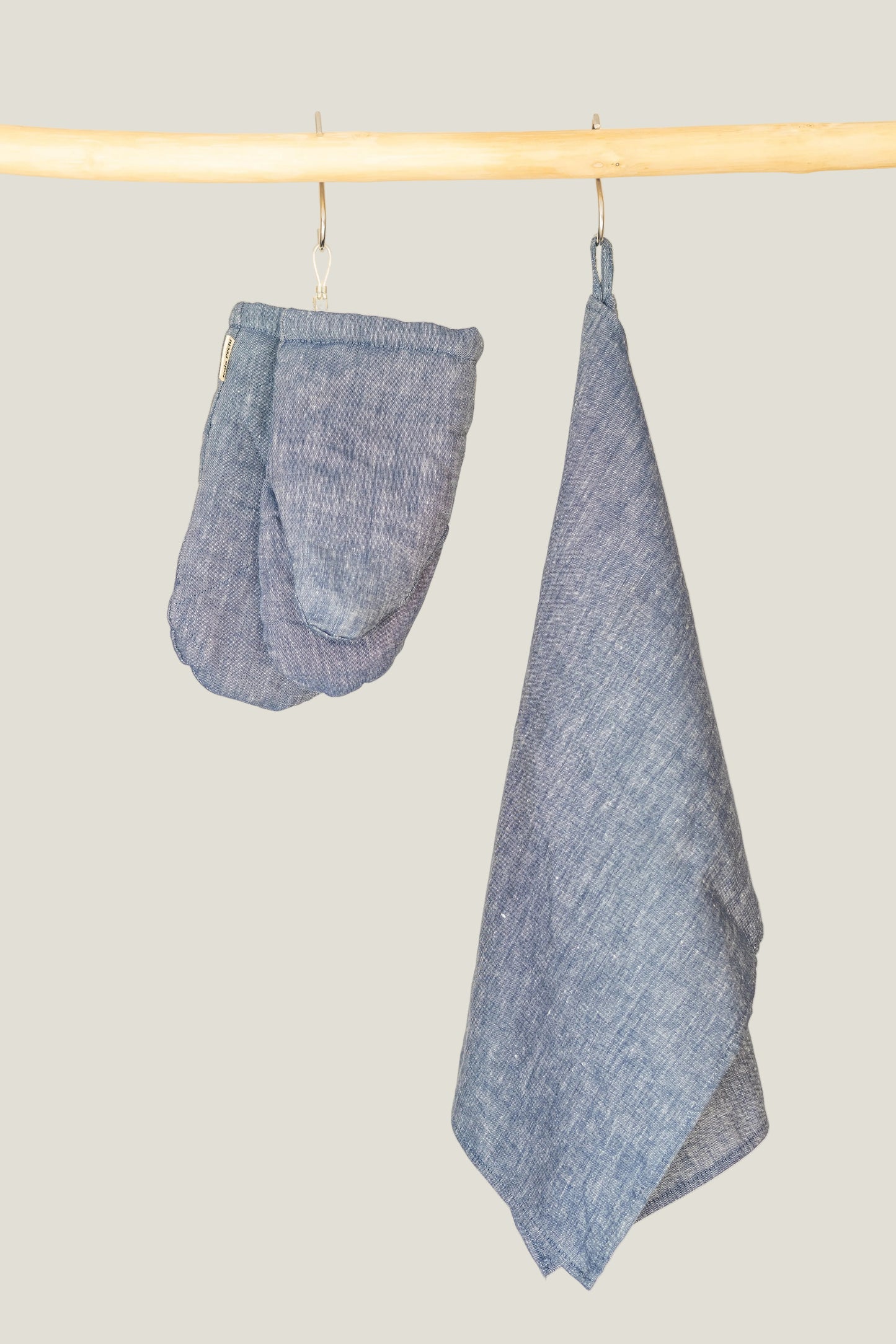 A blue linen  tea towel hanging from a metal hook on a wooden rod next to a matching fabric pot holder set against a plain cream background.