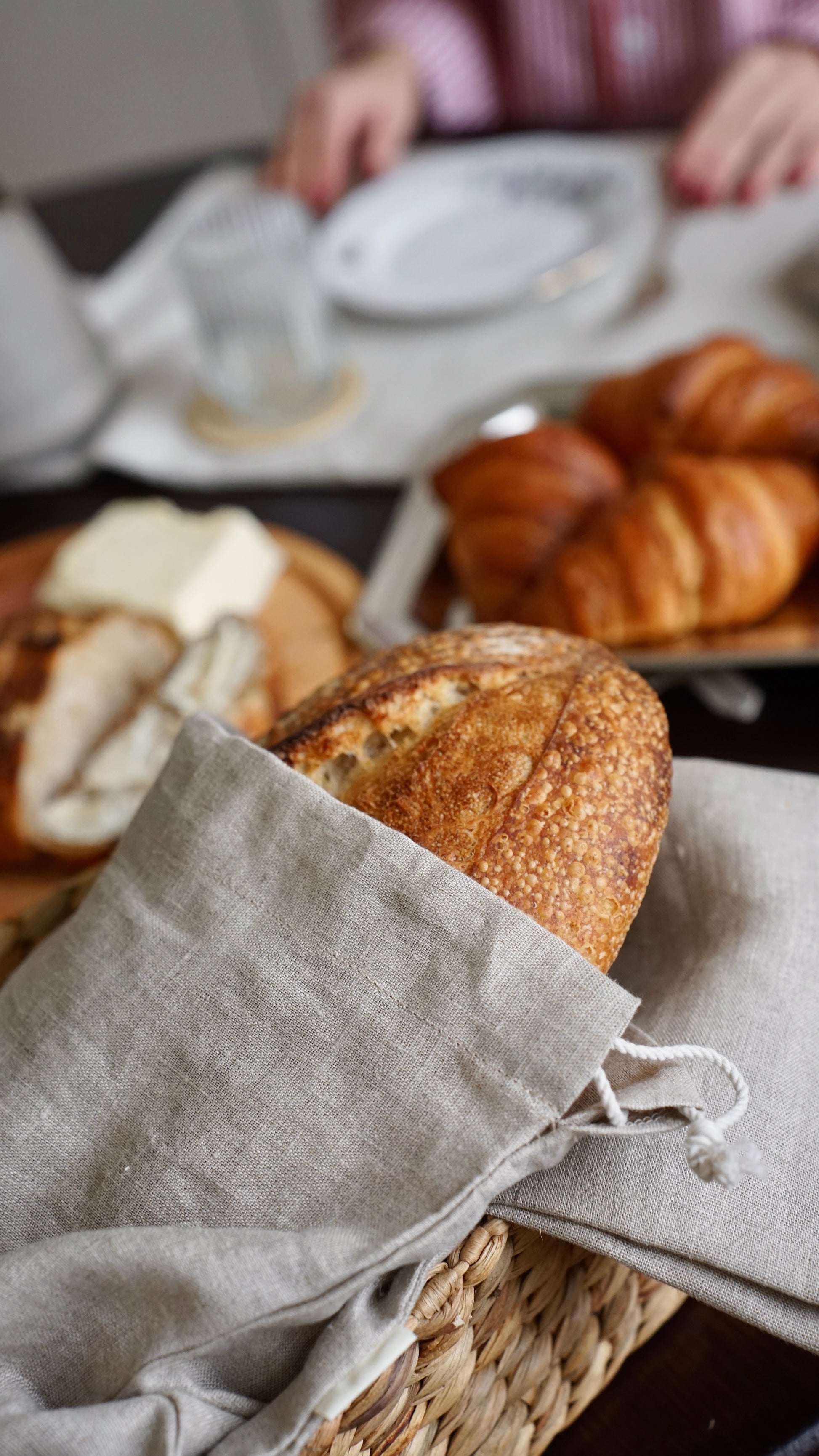 A close-up of a rustic round loaf of bread resting in a woven basket lined with a light beige linen drawstring bag, with other breads and a breakfast table setting blurred in the background.