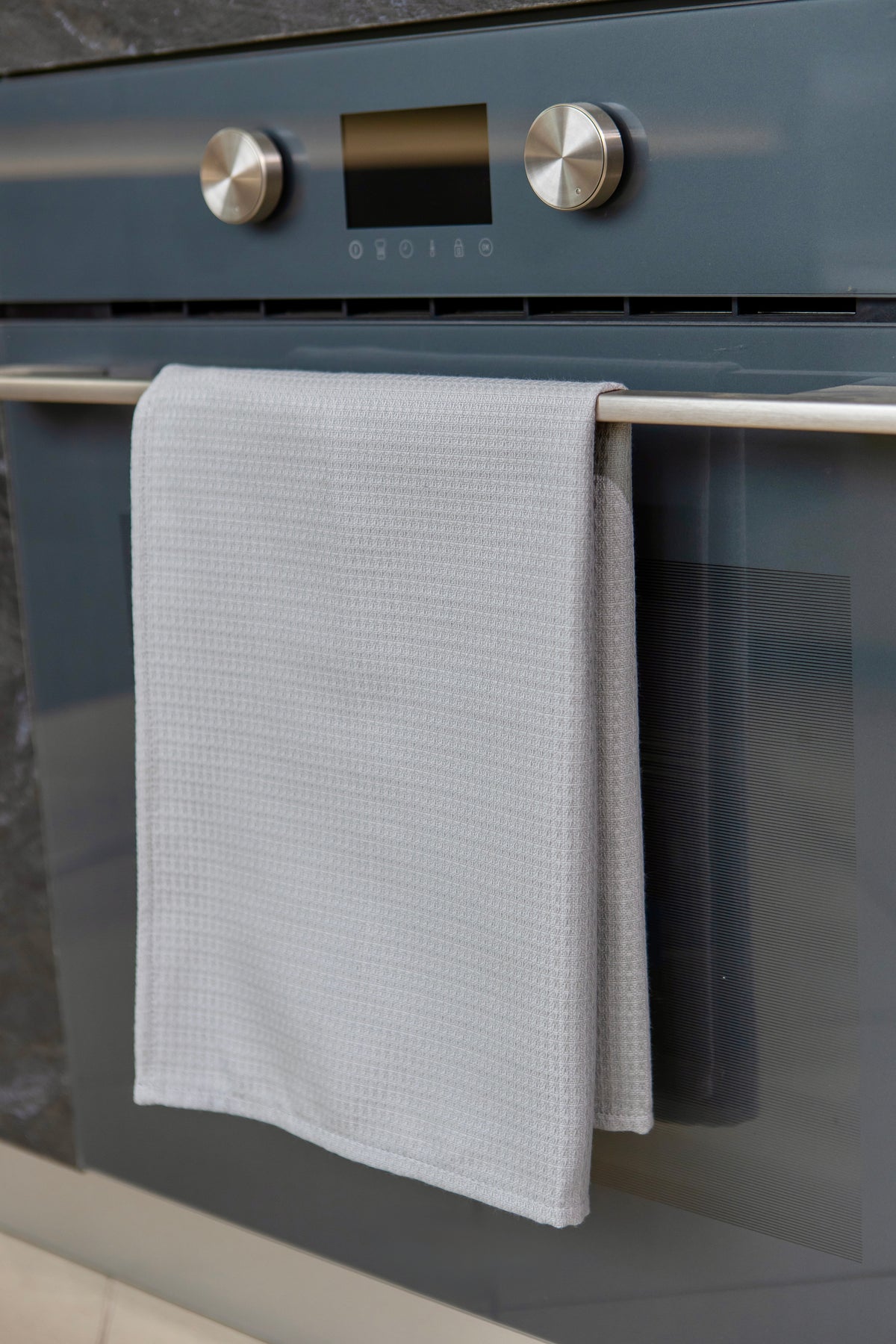 A light gray Kolo Pechi waffle-weave kitchen towel from the Lviv Sky collection hanging from the handle of a dark slate oven.
